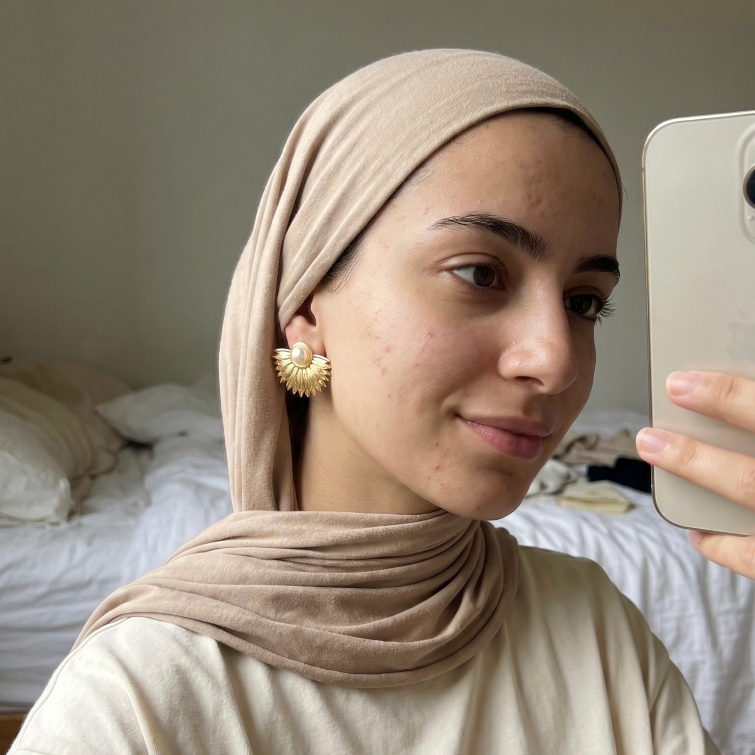 Woman wearing a beige hijab taking a mirror selfie in a bedroom.