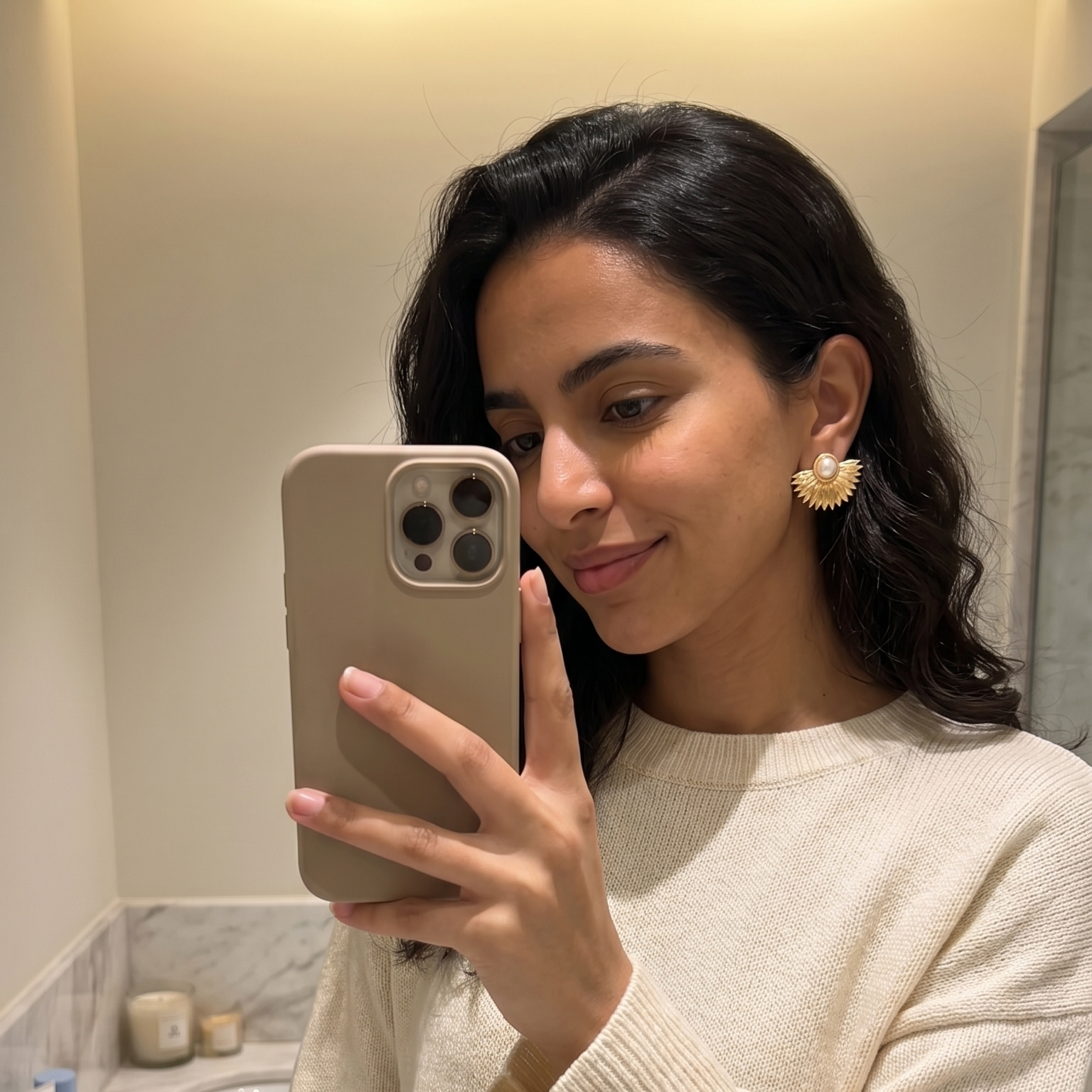 Woman taking a mirror selfie in a bathroom wearing a cream sweater.
